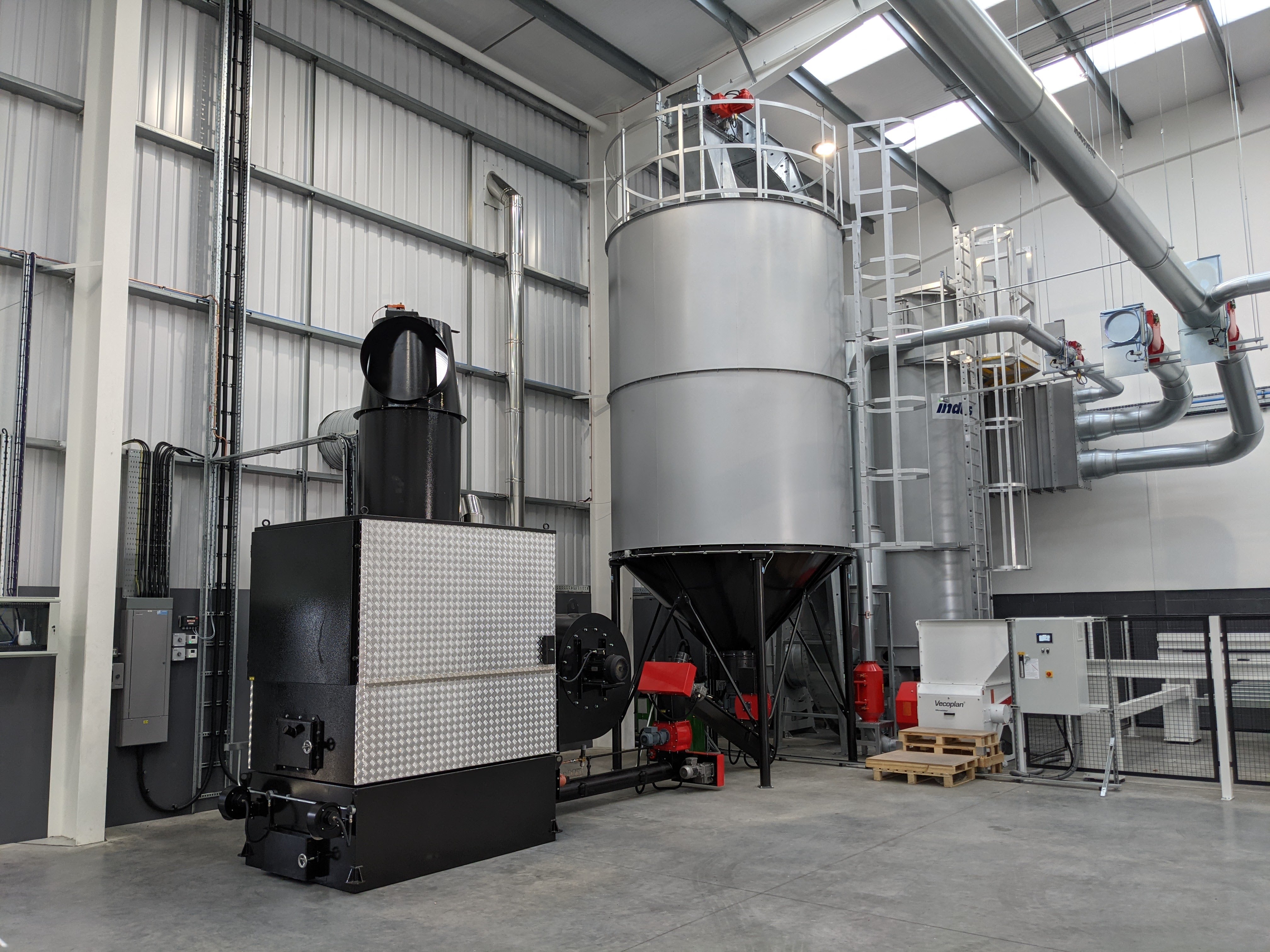 More front-on view of WA 300 warm air heater with C28 silo fed by drag conveyor in a panel processing factory, showing the dust extraction system more clearly and chipper visible